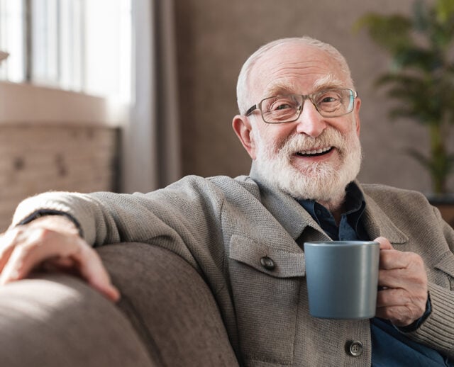 A person with glasses and a beard smiling while holding a mug, sitting on a couch in a cozy, well-lit room.