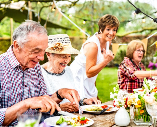 Three people enjoying a meal outdoors, surrounded by flowers and string lights, exuding warmth and happiness in a garden setting.