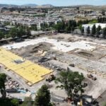 Aerial view of a construction site surrounded by suburban homes, trees, and distant hills under a clear, blue sky.