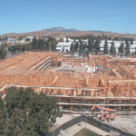 A large wooden construction project surrounded by trees and distant hills under a clear sky, with several people visible on site.