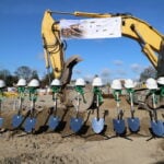 Shovels with helmets and bows stand before a large excavator at a construction site under a clear blue sky.