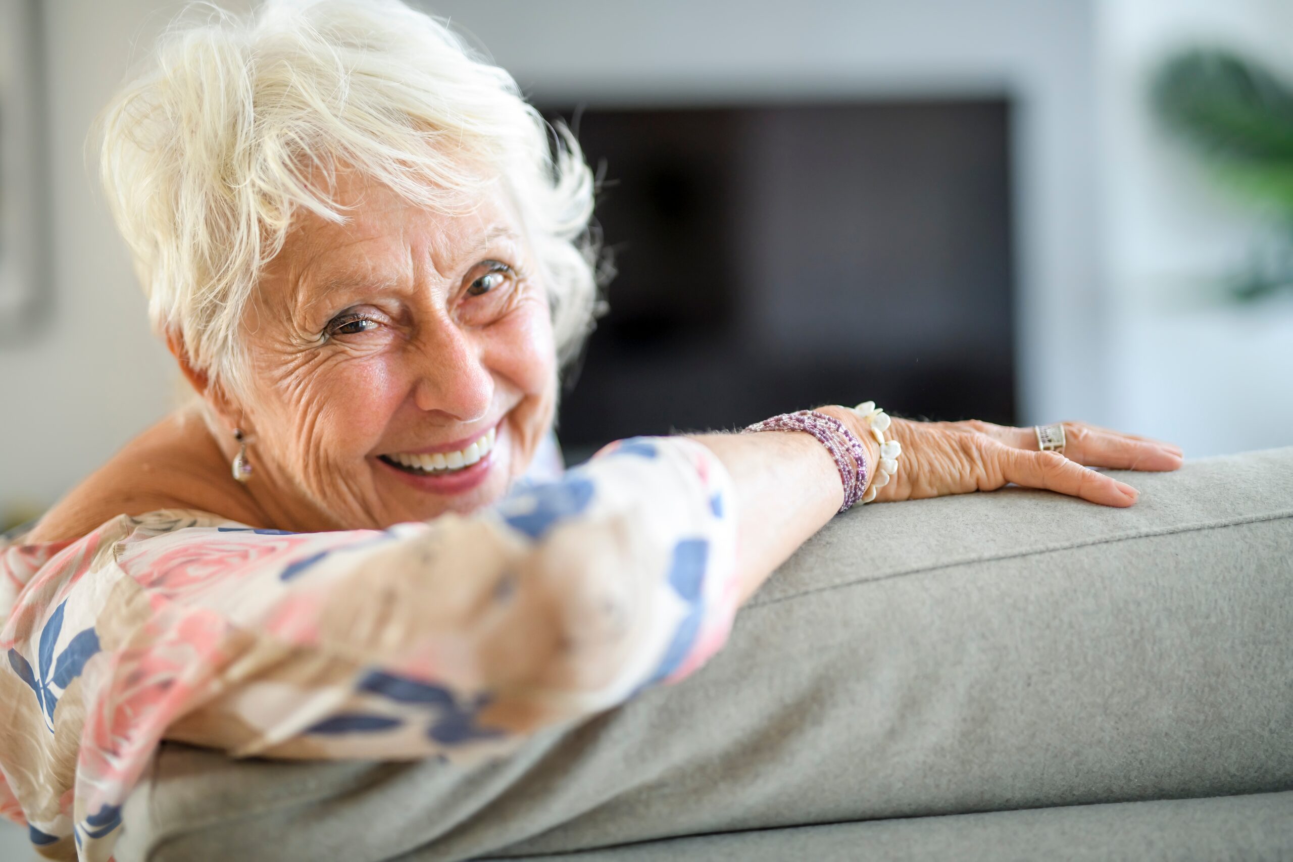 A smiling person with white hair leans on a sofa, wearing patterned clothing and bracelets, in a cozy, softly lit room.