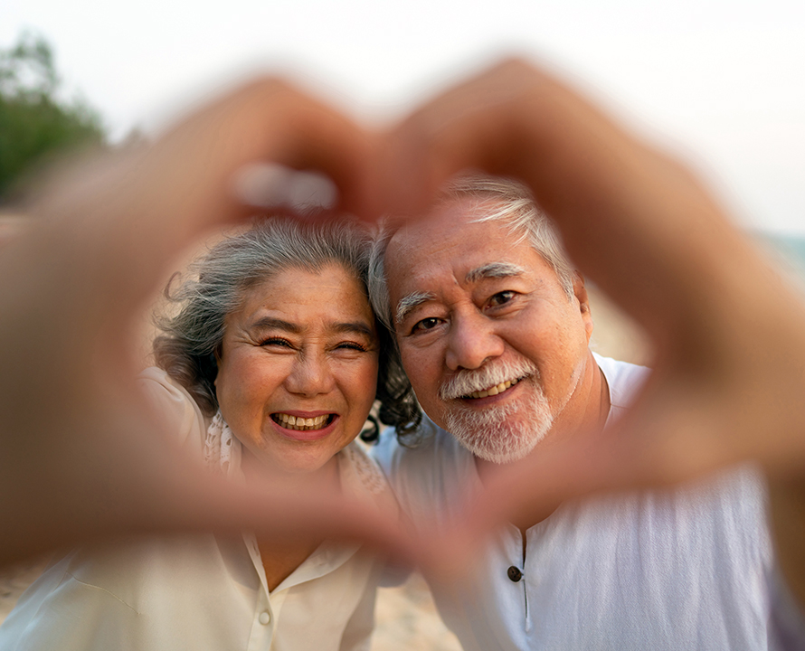 Two smiling people framed by hands forming a heart shape, outdoors, with green trees and sky in the background.