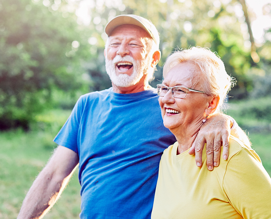 Two people are laughing together outdoors in sunny weather, with lush greenery in the background, expressing joy and companionship.
