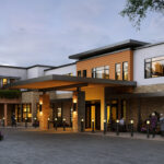 Modern building with large windows, outdoor seating, and warm lighting. Two people converse near entrance, surrounded by trees and landscaped flowers. Evening setting.