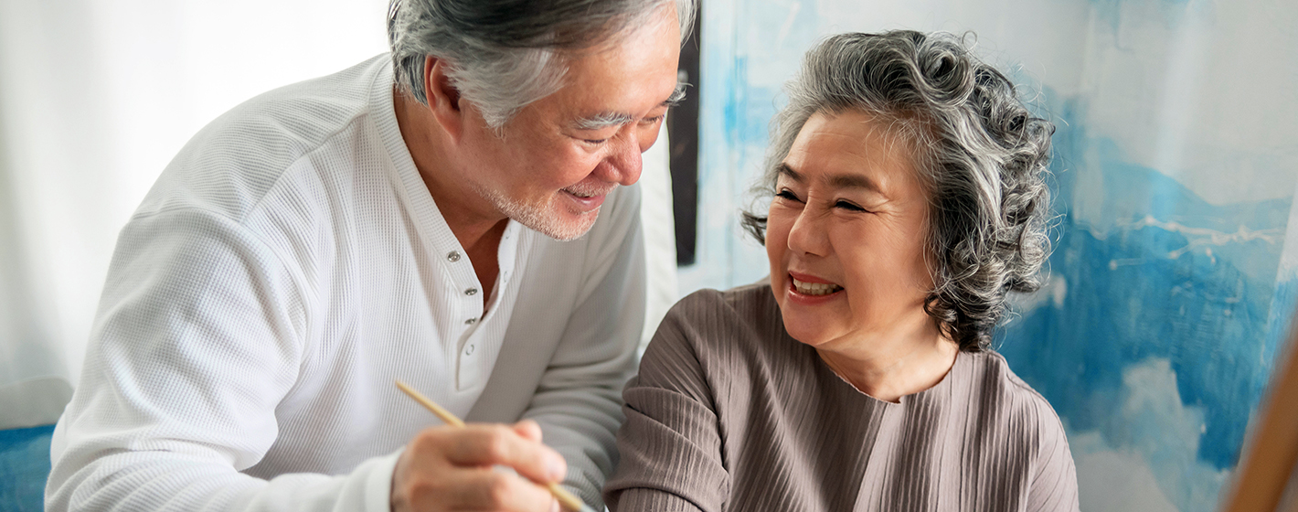 Two people smiling together indoors, one holding a paintbrush. There's a colorful, abstract blue painting in the background, creating a joyful atmosphere.