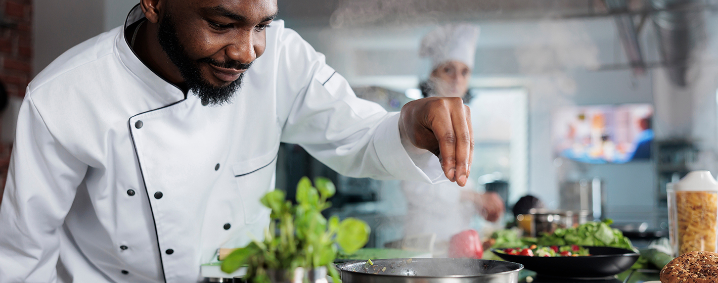 Two chefs in a professional kitchen, one seasoning a dish with fresh ingredients, wearing white uniforms. Background blurred with culinary tools visible.