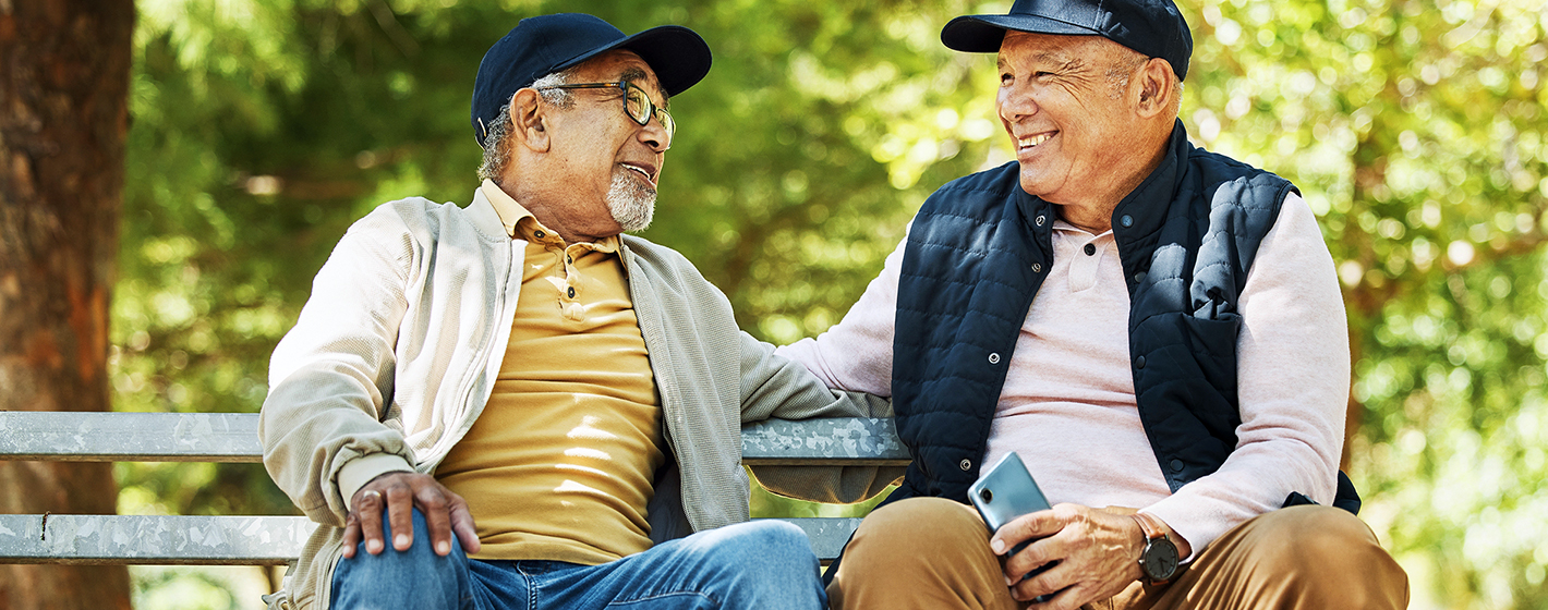 Two people sitting on a park bench, smiling and conversing, amidst a backdrop of lush green trees.