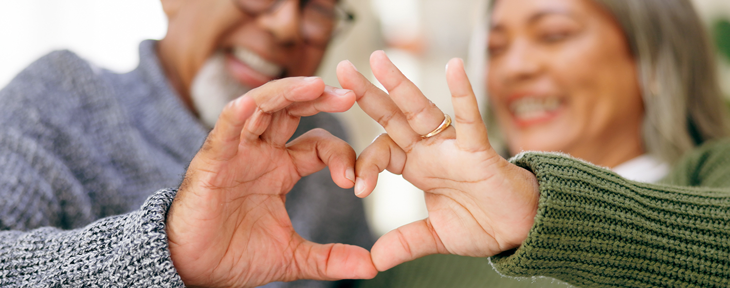 Two smiling people create a heart shape with their hands, signifying love and connection, wearing cozy sweaters, with a blurred background.