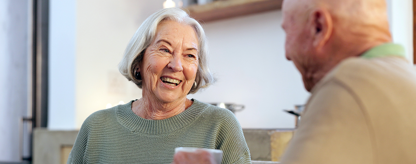 Two people are smiling and talking indoors, creating a warm, friendly atmosphere. The background shows minimalistic decor with shelves and soft lighting.