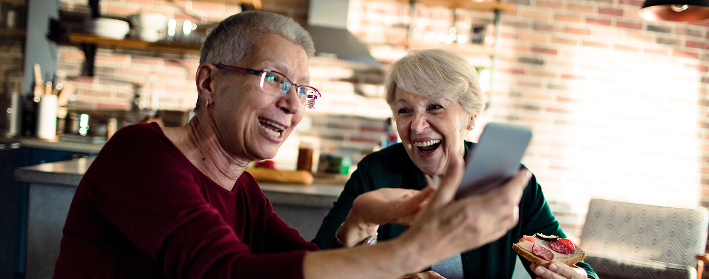 Two persons smiling and taking a selfie in a cozy kitchen, with a brick wall background and one holding a sandwich.
