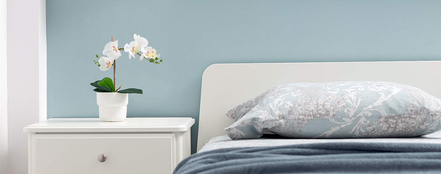 Bedroom scene with a white nightstand, potted orchid, and textured pillow on a bed against a soft blue wall. Simple and serene.