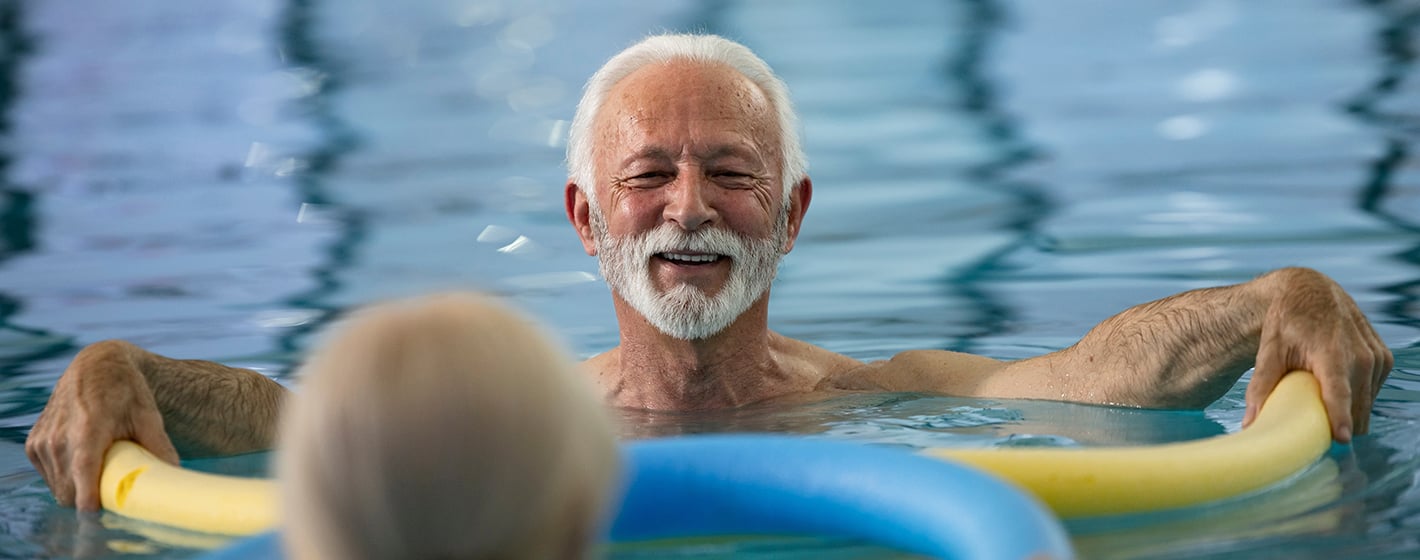 Two people enjoy a swimming session, one smiling while holding a pool noodle. The setting is an indoor pool with clear water.