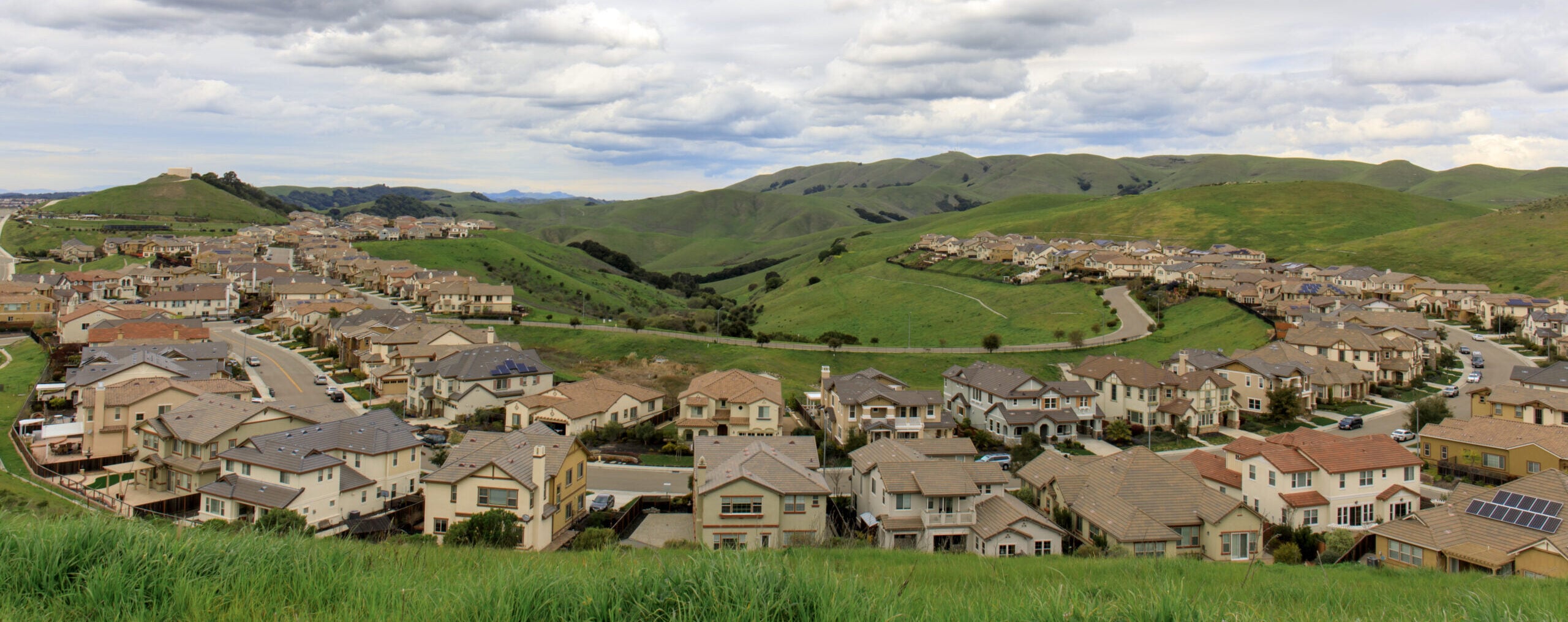 A suburban neighborhood is nestled in rolling green hills under a cloudy sky, featuring rows of modern homes and winding roads.