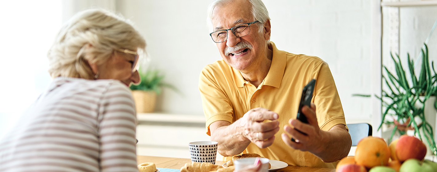 Two people sit at a table with fruit and pastries, smiling and looking at a smartphone.
