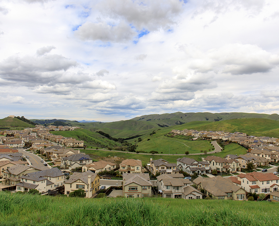 A suburban neighborhood nestled among rolling green hills under a partly cloudy sky; no recognizable landmarks or historical buildings are visible.