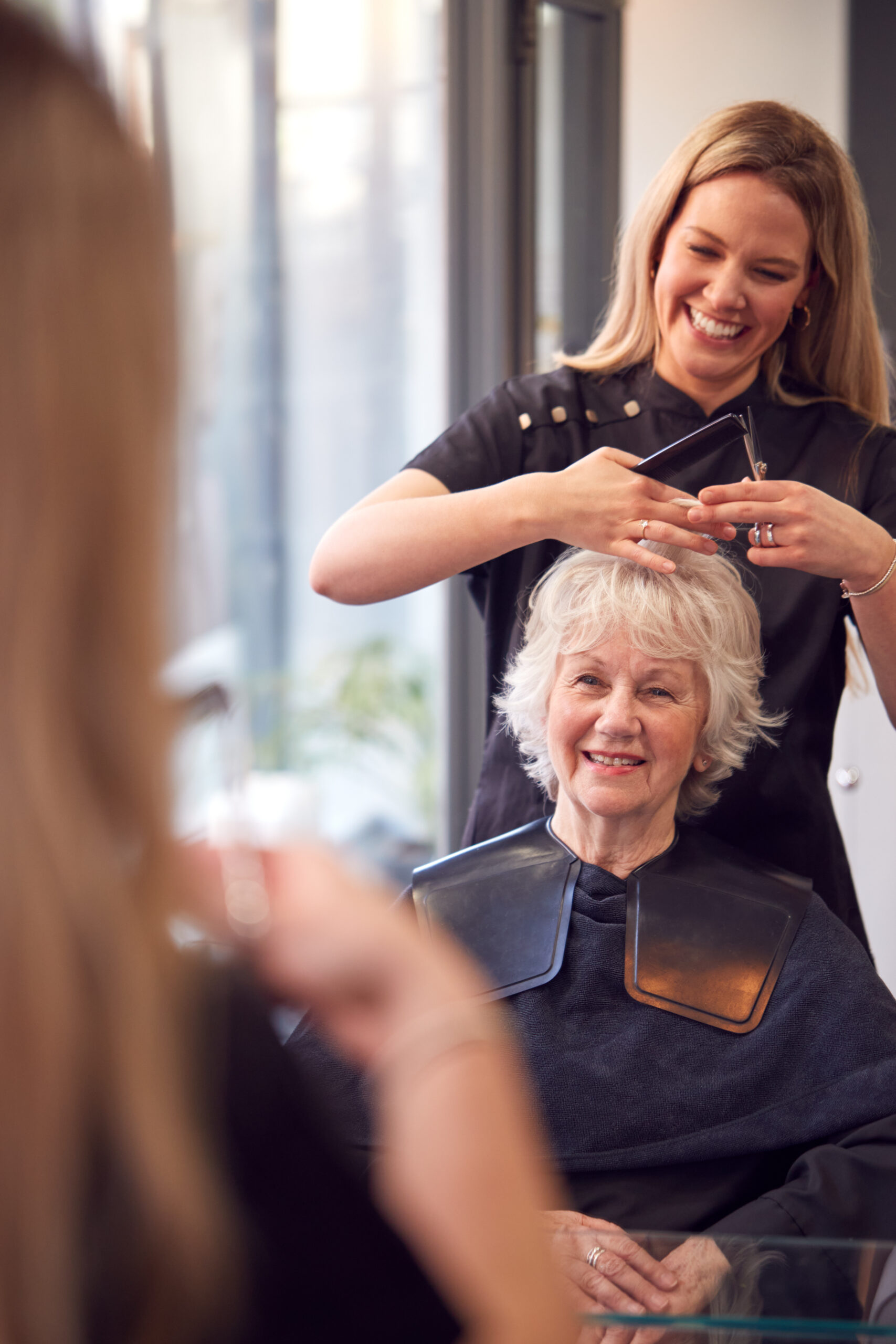 Two people in a hairstyling salon; one styling the other's hair while they both smile, reflected in a mirror.