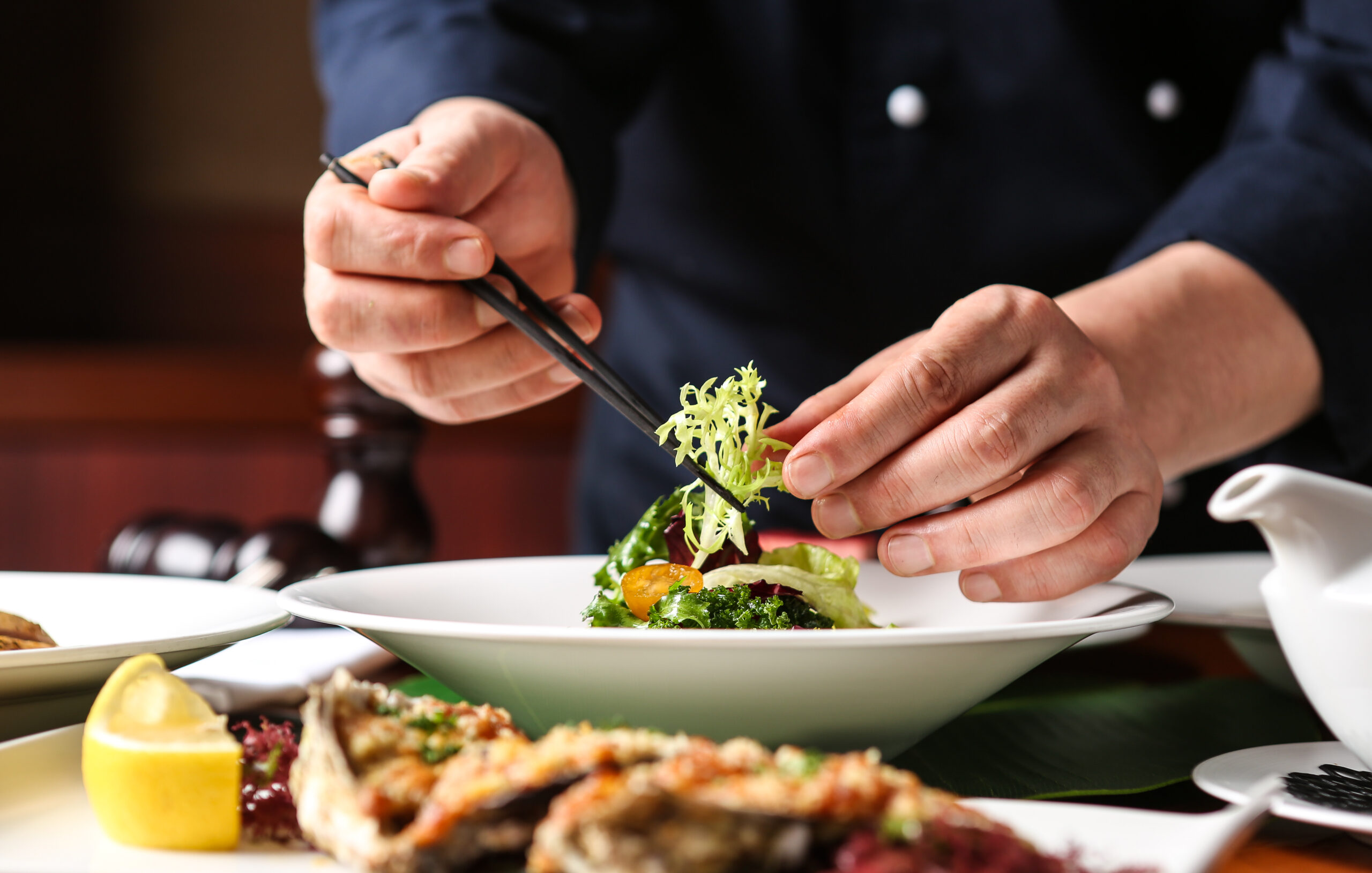 A person in a dark outfit artfully arranges a plated salad with chopsticks, surrounded by other elegantly presented dishes on a dining table.
