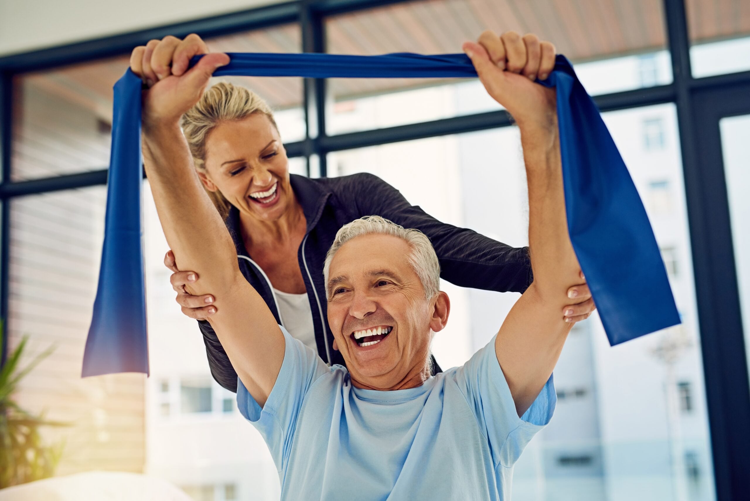 Two people smiling, exercise together indoors. One assists the other with a blue resistance band. Bright, modern room background enhances energetic atmosphere.