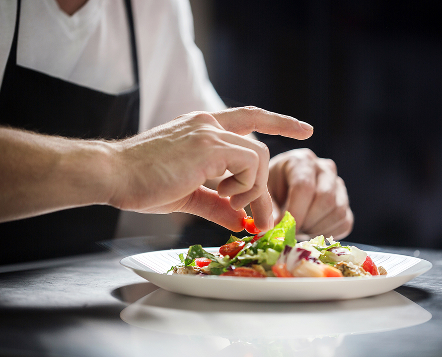 A person carefully garnishes a fresh salad on a white plate, focusing on presentation with vibrant vegetables and greens, under soft lighting.