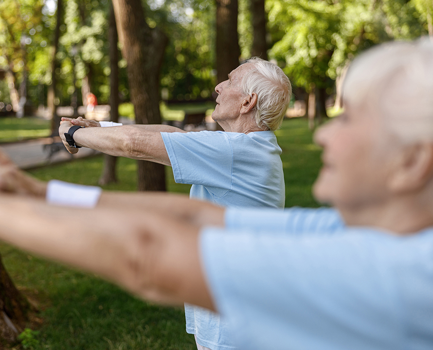 Two people in a park, engaged in outdoor exercise, surrounded by lush green trees. Both wear light blue shirts.