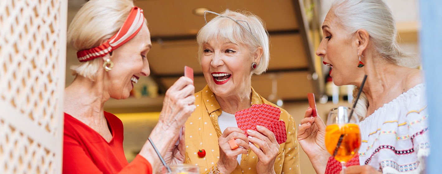 Three people are joyfully playing cards, holding drinks, and laughing together in a cozy indoor setting.