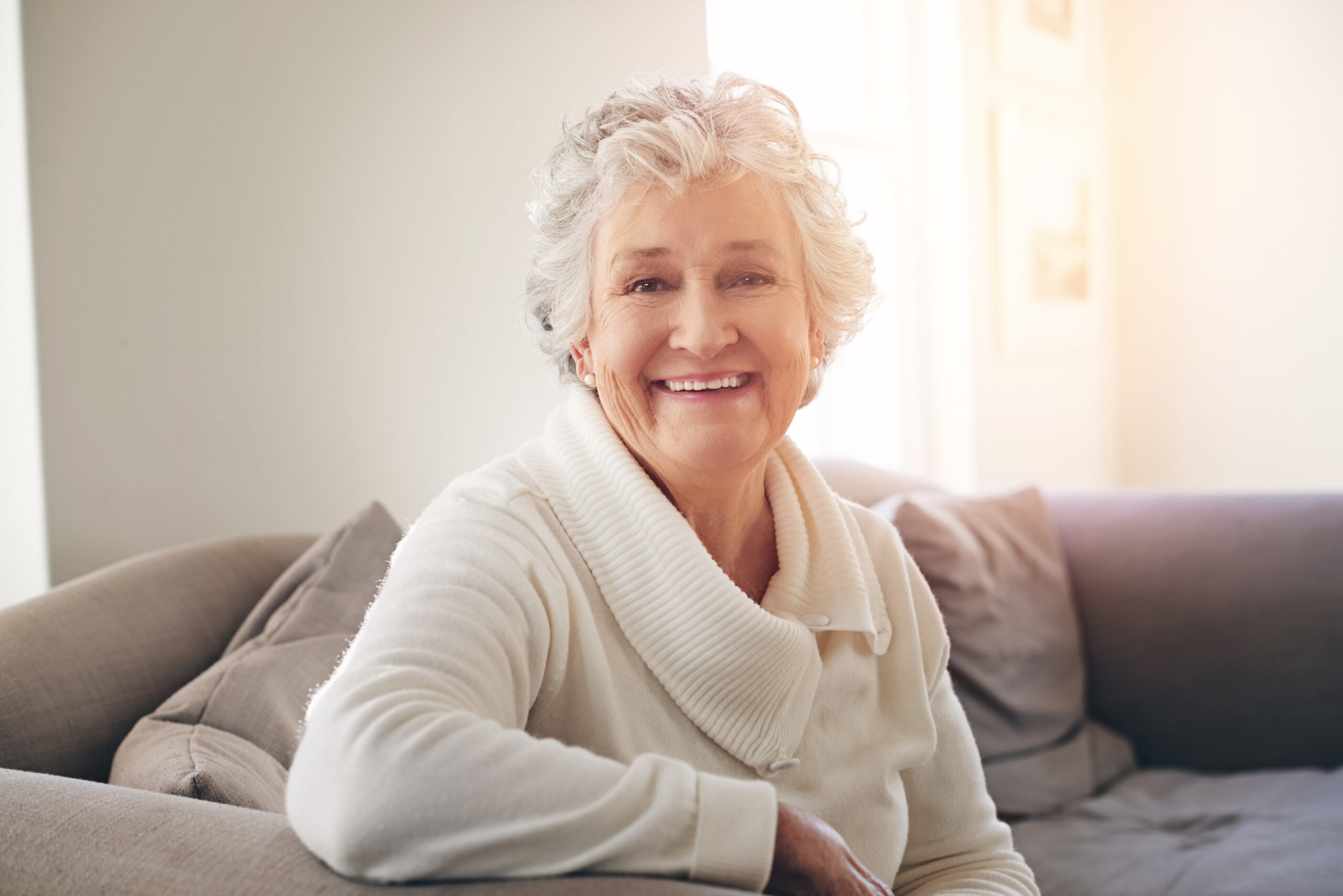 A smiling person with short gray hair and a white sweater sits on a couch in a softly lit room.