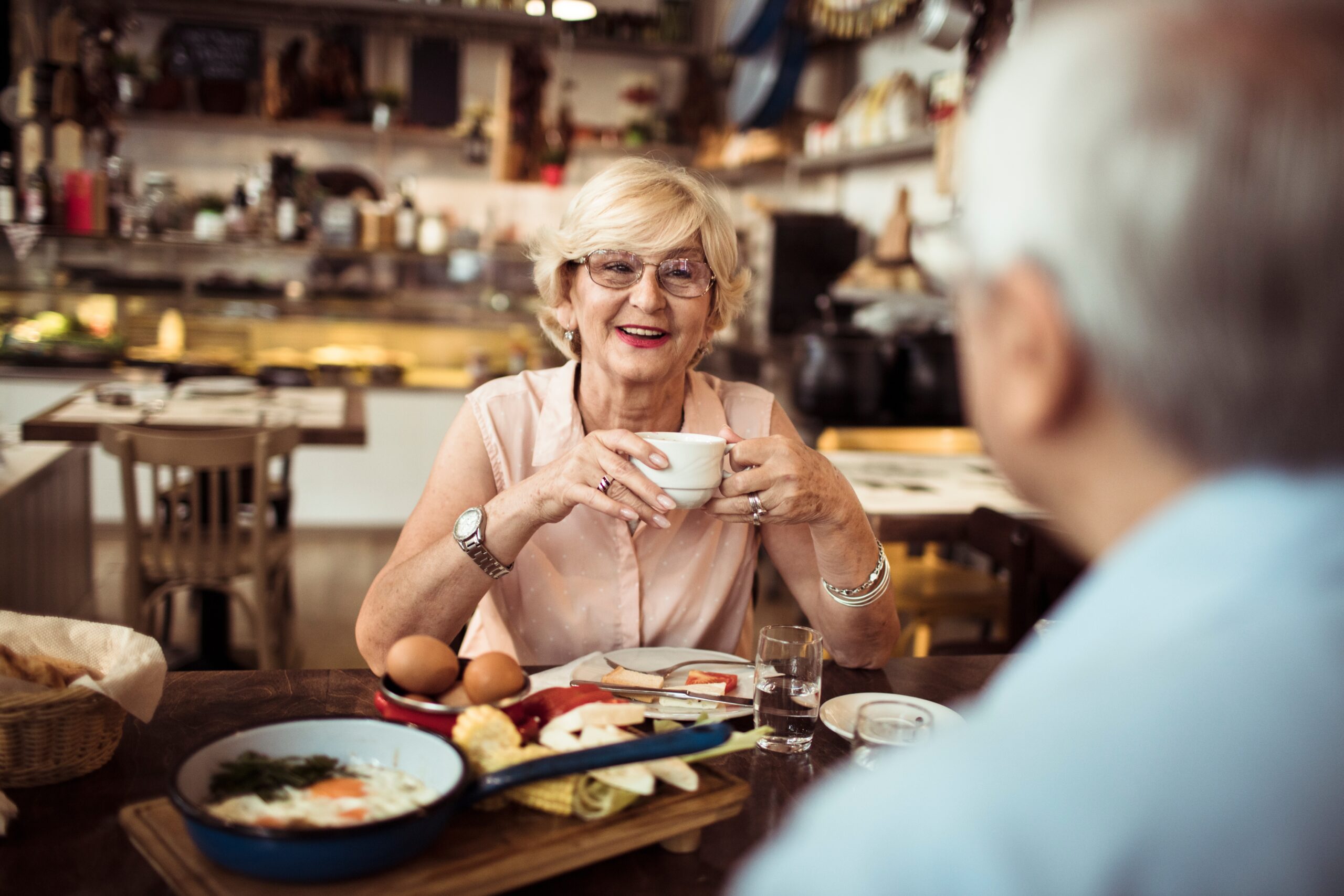 Two people enjoying breakfast in a cozy café. One person holds a cup, smiling, with a plate of food on the table.